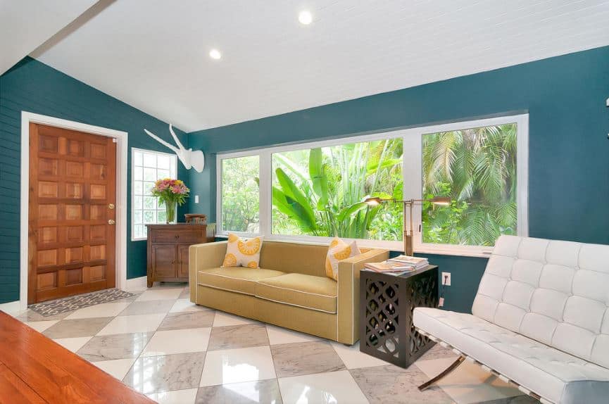 这间热带风格的门厅有着美丽的绿色墙壁，与窗外郁郁葱葱的绿色热带植物相匹配，窗外是舒适的米色沙发，在白色和灰色的格子地板和红木正门的映衬下显得格外突出。