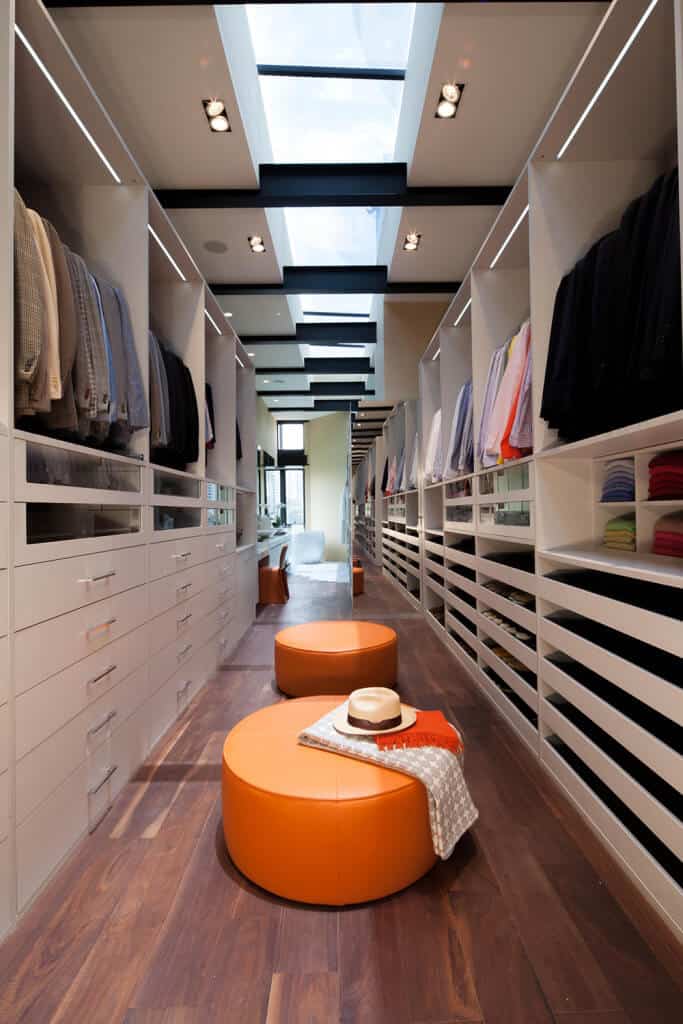 橙色皮革座椅为这个狭长的步入式衣柜带来了色彩的流行，展示了宽大的木板地板和常规的白色天花板，天花板上装有嵌入式灯和贯穿整个房间的天窗。