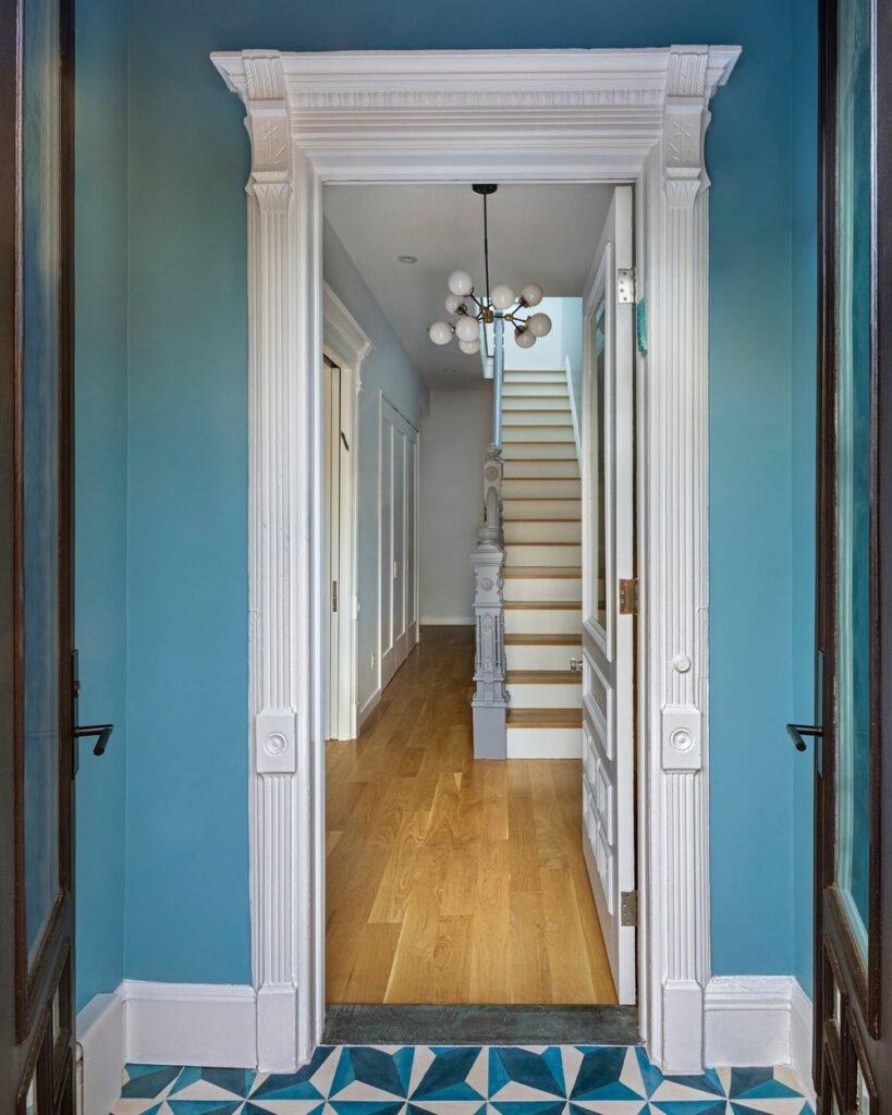 这个简单的维多利亚风格的门厅从蓝色图案的地砖过渡到硬木，因为它移动到第二个门口，有一个优雅的白色框架，木楼梯的底部平台，顶部有一个现代装饰吊灯。