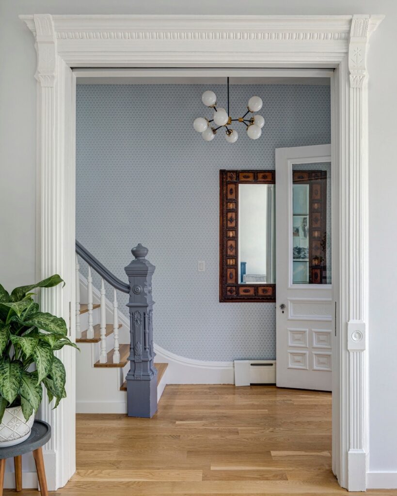 这是维多利亚风格的门厅的另一个角度，你可以看到楼梯的底部平台装饰着一面壁挂式镜子，镜子有一个优雅的木制框架，使它在有图案的浅蓝色墙纸的衬托下脱颖而出。
