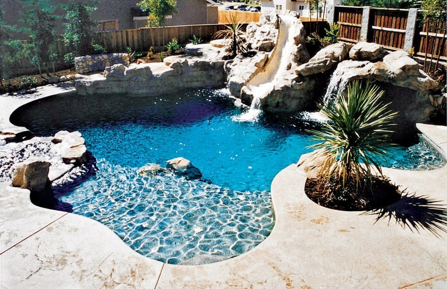 这个后院游泳池融合了瀑布的特征，并带有大石头和微妙的植物的幻灯片。 