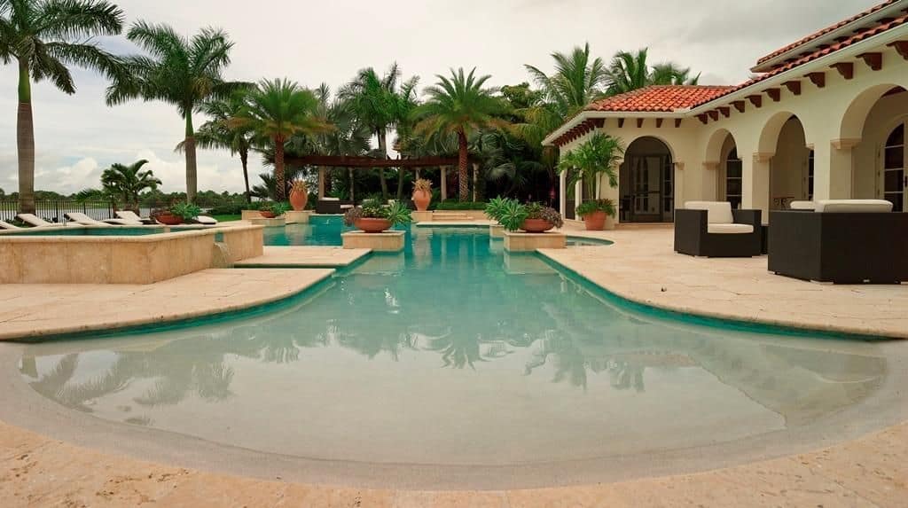 地中海房屋，有一个海滩入口游泳池，周围是高耸的棕榈树。它提供凉棚和大量白色缓冲躺椅。