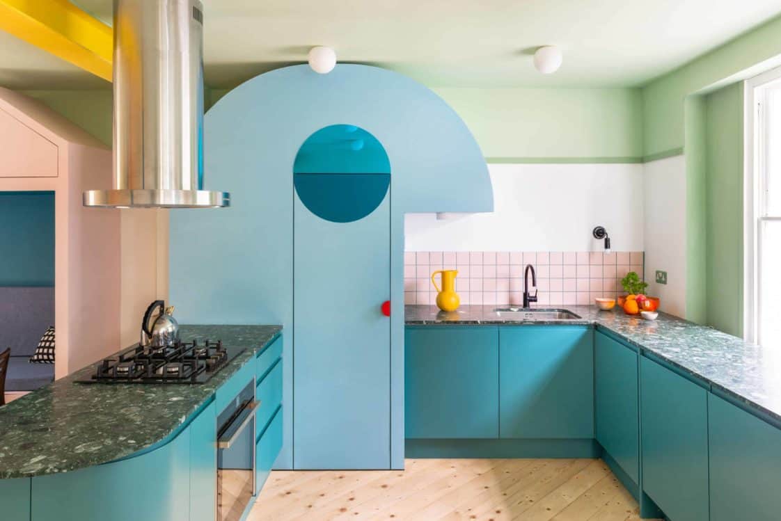这是一个不拘一格的厨房，蓝绿色的橱柜，半岛和粉红色的地铁瓷砖在后溅。冰箱的色调也和橱柜一样。