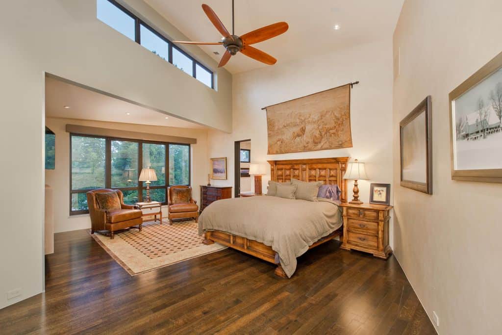 这间主卧室设有玻璃镶板窗边的休息区，以及一张由林地挂毯突出的舒适木床。它有米黄色的墙壁，高高的天花板上安装着青铜风扇和嵌入式灯。