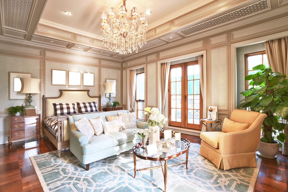 豪华主卧室拥有一个舒适的床和一个休息区在蓝色图案的地毯由一个神话般的水晶吊灯点燃。它有深色硬木地板和棕色护墙板墙延伸到华丽的托盘天花板。