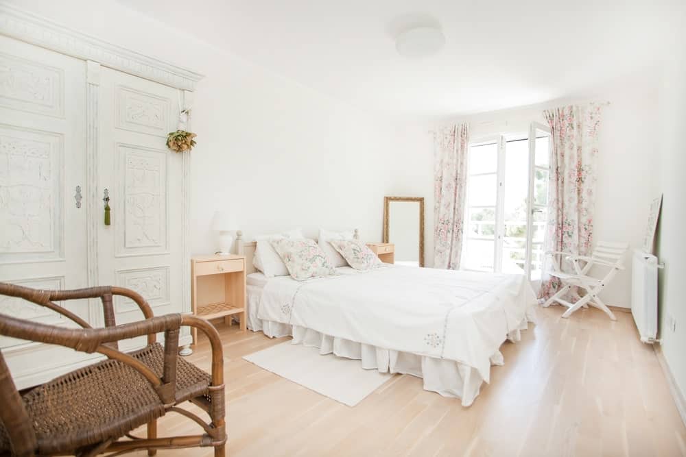 明亮的主卧室，浅色硬木地板和一扇覆盖着花卉窗帘的法式门。它拥有一张有裙子的床和一个迷人的白色衣柜，设计有复杂的细节。