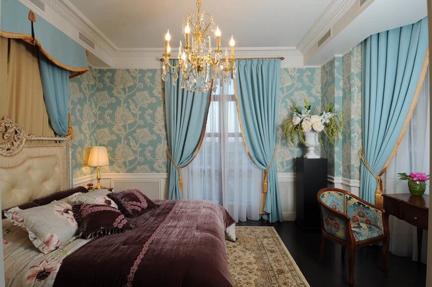 这间经典的主卧室覆盖着白色护墙板和花卉壁纸，与天蓝色的窗帘相辅相成。一张优雅的簇绒床和一盏别致的水晶吊灯为房间增添了优雅。
