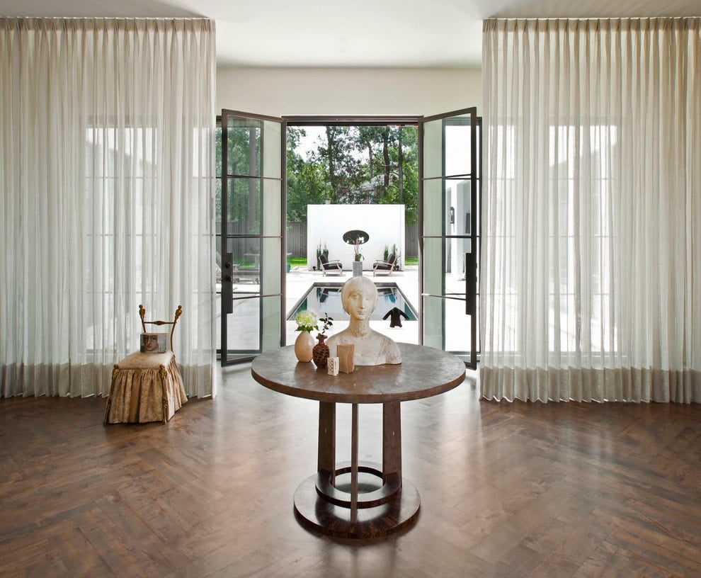 经典的门厅，法式入口门窗覆盖着透明的窗帘。里面有一把带裙边的椅子和一张中间的圆桌，上面有人字形木地板上的半身像雕塑。