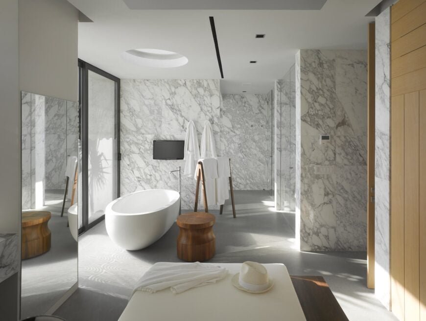 灰色大理石墙壁贯穿了这间主浴室，设有步入式淋浴间和圆形天窗下的独立浴缸。木制毛巾架和凳子，以及横跨全身镜子的白色软脚凳。