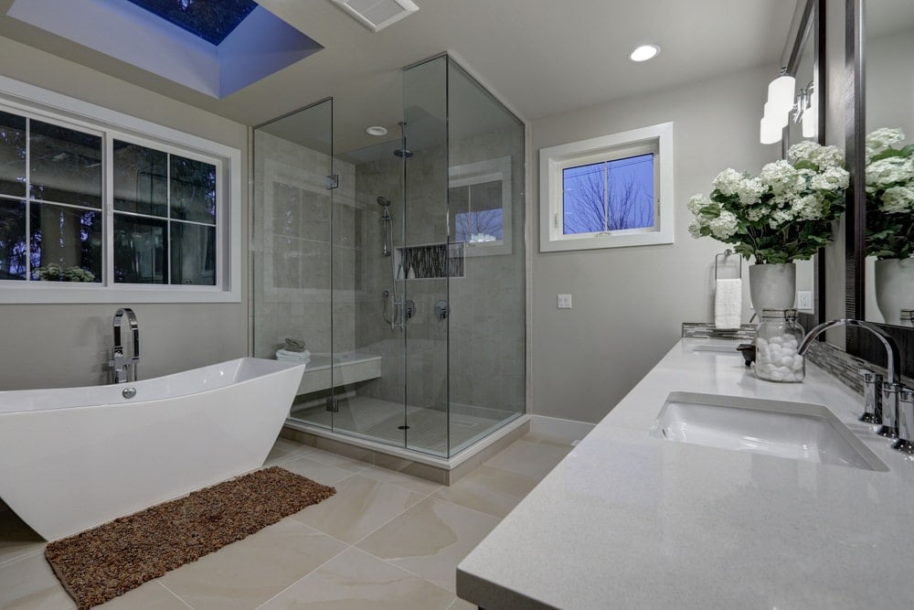 白色框窗和天窗下的独立浴缸与棕色蓬松地毯相映成色。它还配有一间步入式淋浴间和一个双洗手池梳妆台，靠在浅灰色的墙壁上。