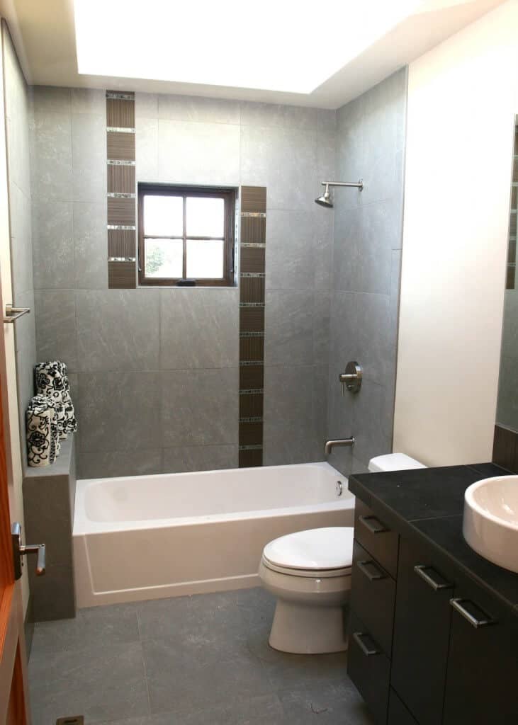自然光从这间灰色浴室的天窗射进来，浴室里有淋浴和浴缸，还有厕所和一个带水槽的深色木质梳妆台。