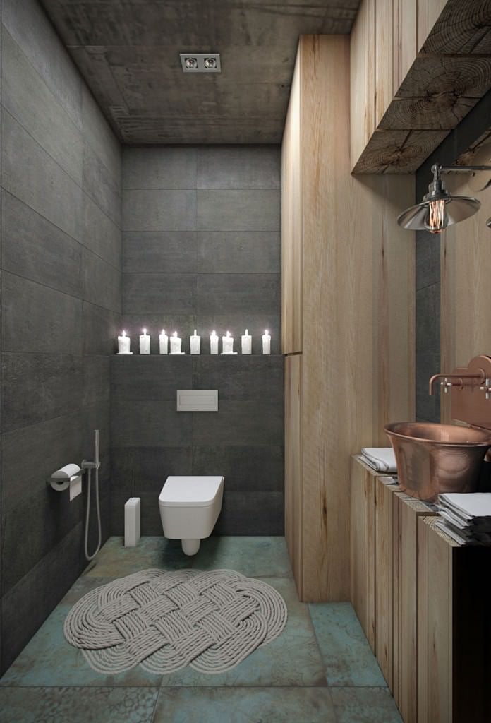 小浴室有灰色的瓷砖墙和破旧的绿色地板，上面有一个可爱的编织地毯。里面有一个挂在墙上的厕所和一个木板梳妆台，梳妆台顶部有一个铜质水槽和配套的夹具。