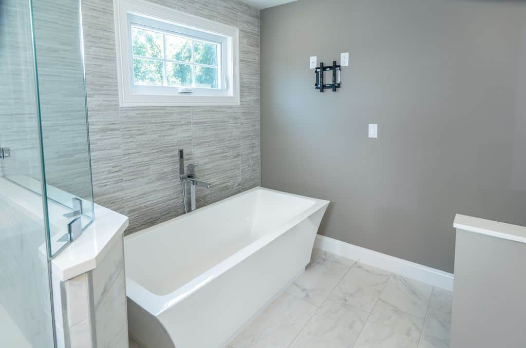 通风的主浴室，大理石瓷砖地板和灰色墙壁，内衬白色底座。里面有一间步入式淋浴间，还有一个独立的浴缸，靠近装有镀铬固定装置的白色框窗。