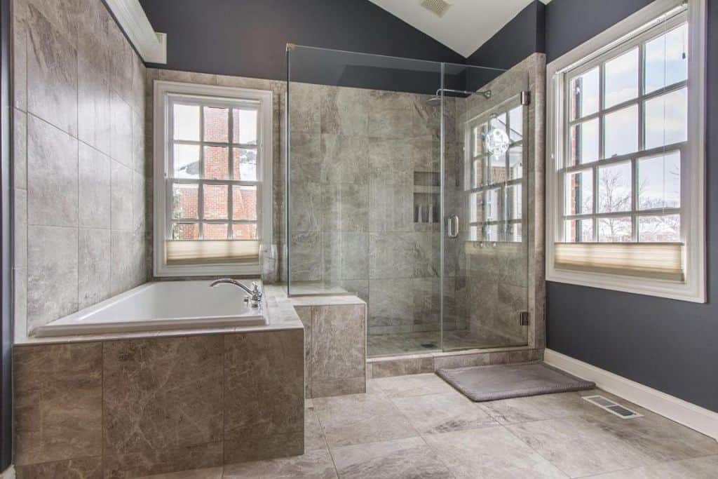 灰色的主浴室带有大教堂天花板和米黄色瓷砖地板，延伸至浴缸周围和后挡板。它有一间步入式淋浴间和白色框窗，能让充足的自然光照进来。