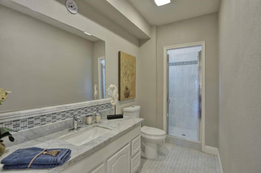 大理石台面的梳妆台配有无框镜子，在白色瓷砖地板上的传统厕所两侧设有步入式淋浴间。它被灰色的墙壁包围，装饰着华丽的花卉艺术作品。