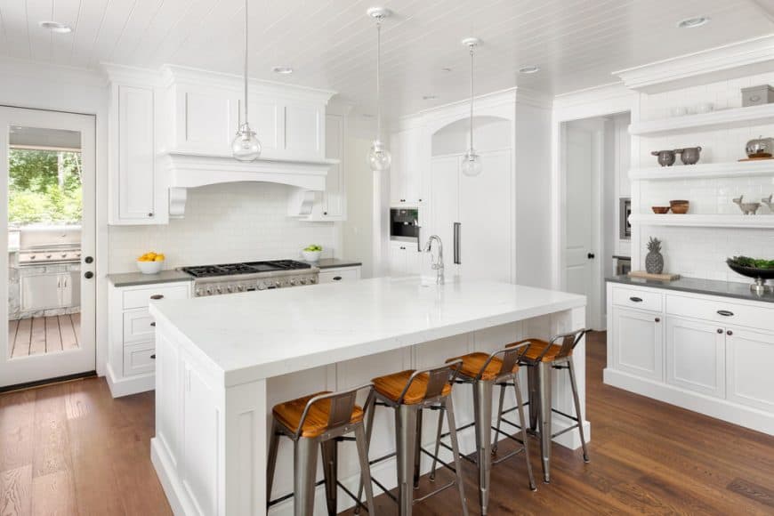 这间明亮的白色厨房的硬木地板与厨房岛台的白色摇酒柜和抽屉以及周围的橱柜很好地融合了白色的船型吊顶。这地板也与早餐吧的现代凳子很好地搭配。