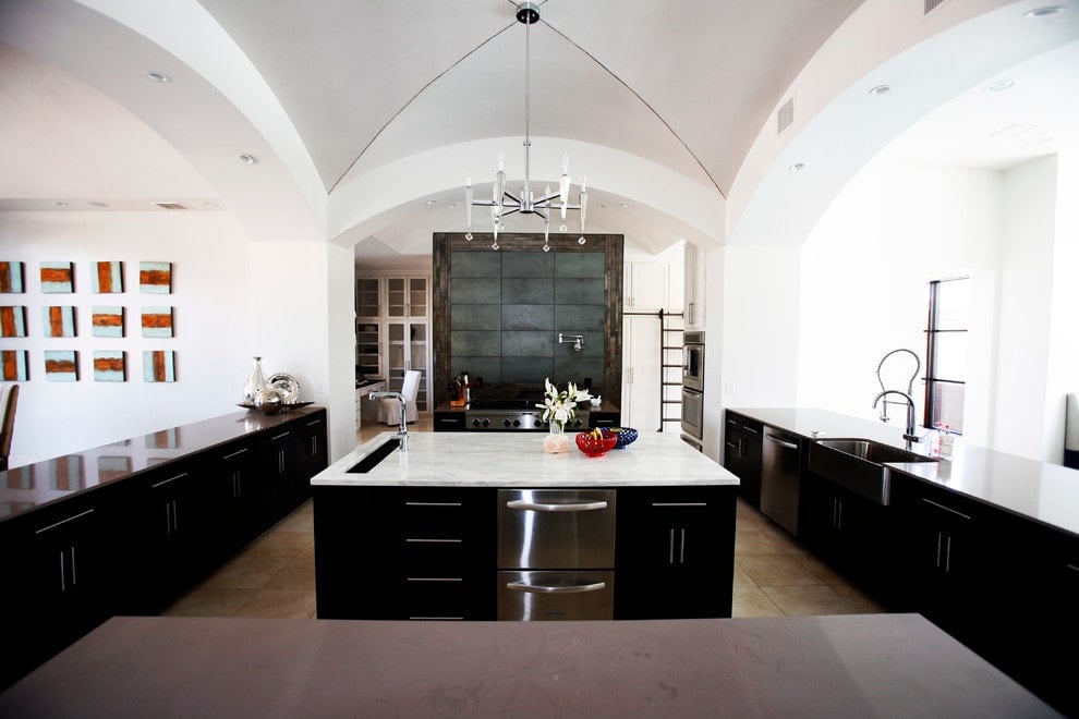 现代厨房，白色墙壁和腹股沟拱顶天花板与黑色橱柜和大理石顶部岛形成对比。它配备了不锈钢用具和搭配铬固定装置的农舍水槽。