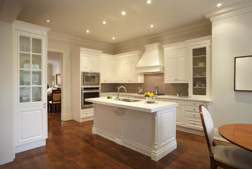 这个优雅厨房明亮狭窄的厨房岛在深色硬木地板的衬托下显得格外突出。这种地板也与明亮的白色摇壶柜和木制结构的抽屉形成对比，墙壁上主要是灰色的后挡板。