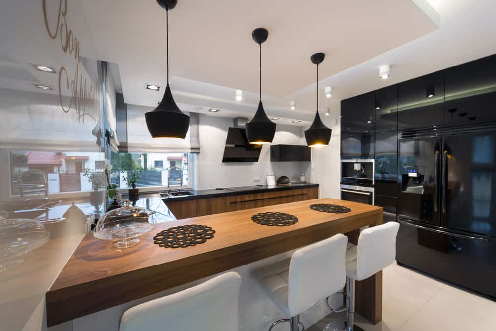 这个现代厨房的黑色家电给它一种精致和奢华的特殊美学。木质早餐吧上方悬挂着白色天花板，黑色吊灯与白色现代凳子形成对比，这进一步增强了这一点。