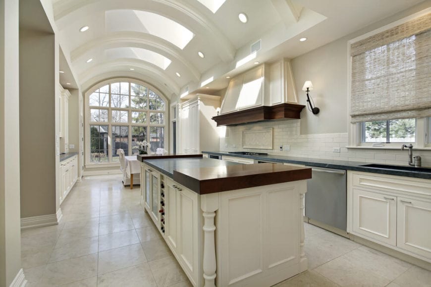 这是一个明亮明亮的厨房，主要是因为它的凹形天花板由大型天窗控制，并通过嵌入式灯光进行增强，形成独特的明亮美学，以补充大型厨房岛的白色摇壶柜和抽屉以及墙壁上的橱柜。