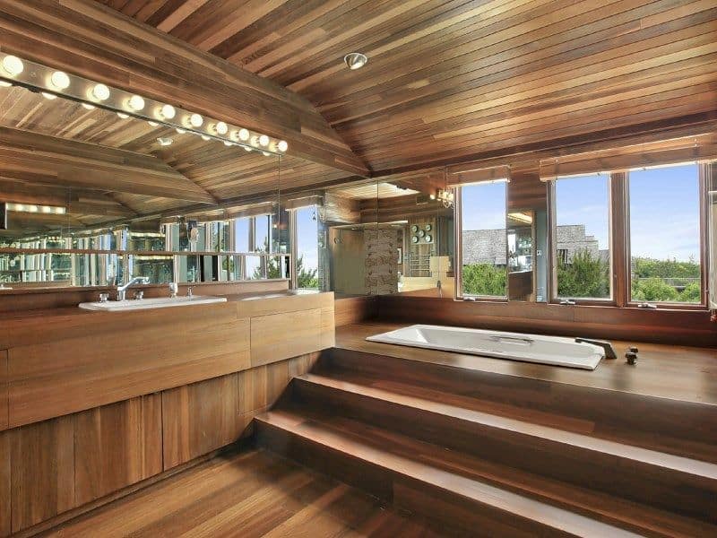 一间现代化的主浴室，拥有时尚的硬木地板、墙壁和木制天花板。它有一个漂浮的梳妆台水槽和一个靠窗的落地式浴缸。