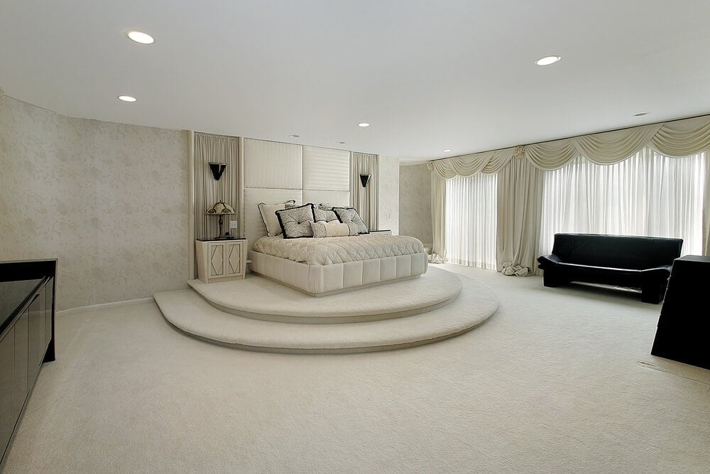 宽敞的主卧室设有两级平台上的白色簇绒床，与地毯地板和米色墙壁融为一体，墙壁上装饰着优雅的窗帘和窗帘。它与黑色橱柜和皮革沙发形成对比，与透明的窗帘形成对比。
