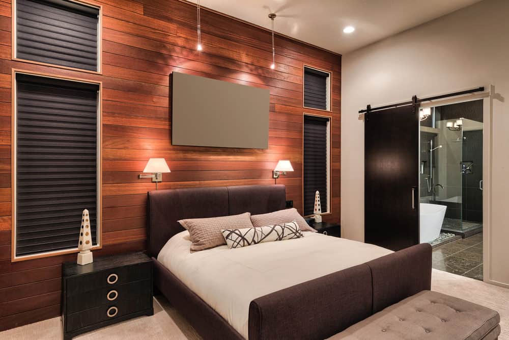 经典的烛台照亮的软垫床两侧黑色床头柜和独特的台灯。这个现代主卧室有米色地毯地板和木板口音壁装有玻璃窗,覆盖着灰色的卷帘。