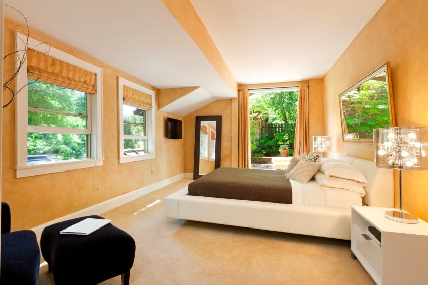 米色地毯地板与主卧室的橙色墙壁融合得很好，白色床头柜和平台床与黑色天鹅绒躺椅形成对比。它由时尚的玻璃台灯照亮，自然的光线从玻璃窗流入室内。