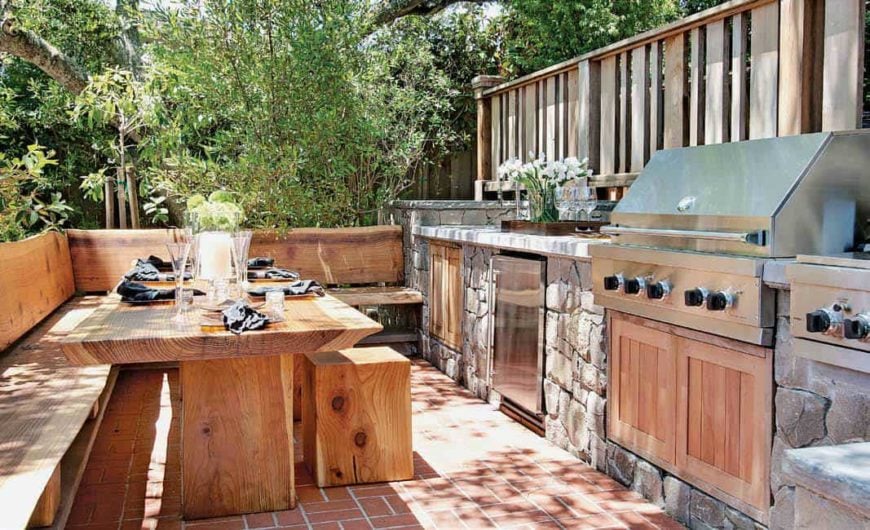 这是一个舒适的户外厨房，有一个裸露的木质餐桌，内置的l形长凳和小凳子，与陶土地板非常相配。这也补充了不锈钢器具的纹理石材结构。