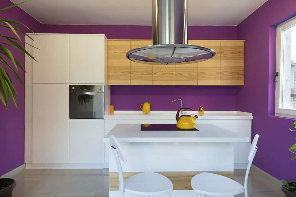 这间小厨房可爱而欢快的紫色墙壁与靠墙的白色现代橱柜相映成趣，水池上方则是裸露的木制浮动橱柜。这是与一个白色的小厨房岛与一对白色现代凳子早餐吧。