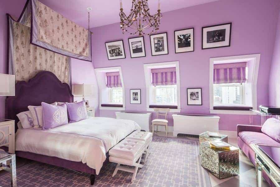 这间紫色的主卧室以一系列黑色相框的照片和挂在白色簇绒脚凳上的华丽串珠吊灯为风格。它展示了一个休息区和一张迷人的软垫床，上面有一个顶篷。