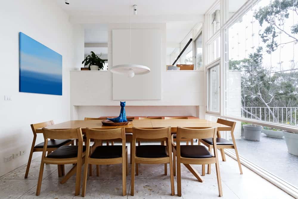 在这个明亮的餐厅里，通高的玻璃创造了一种空间感，瓷砖地板和蓝色帆布画强调的白色墙壁。里面摆满了光滑的软垫椅子和一张木制餐桌，餐桌上挂着白色的吊灯。