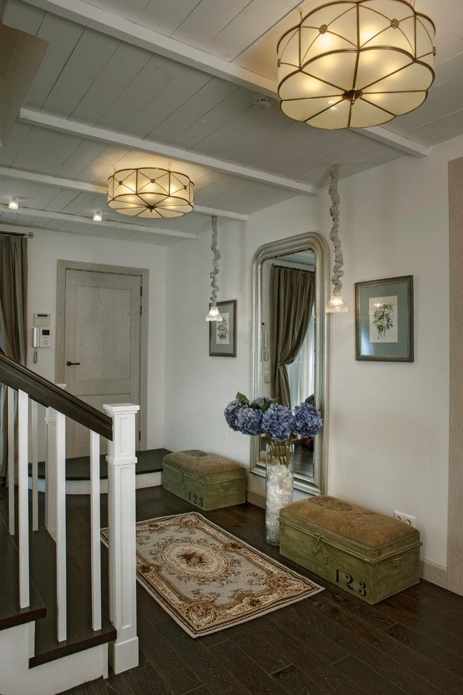 一对储物椅和花卉艺术品在门厅里形成完美的对称，门厅里有一面拱形的全身镜和一块迷人的区域地毯，铺在深色硬木地板上。它由独特的吊坠和可爱的平装灯照明。