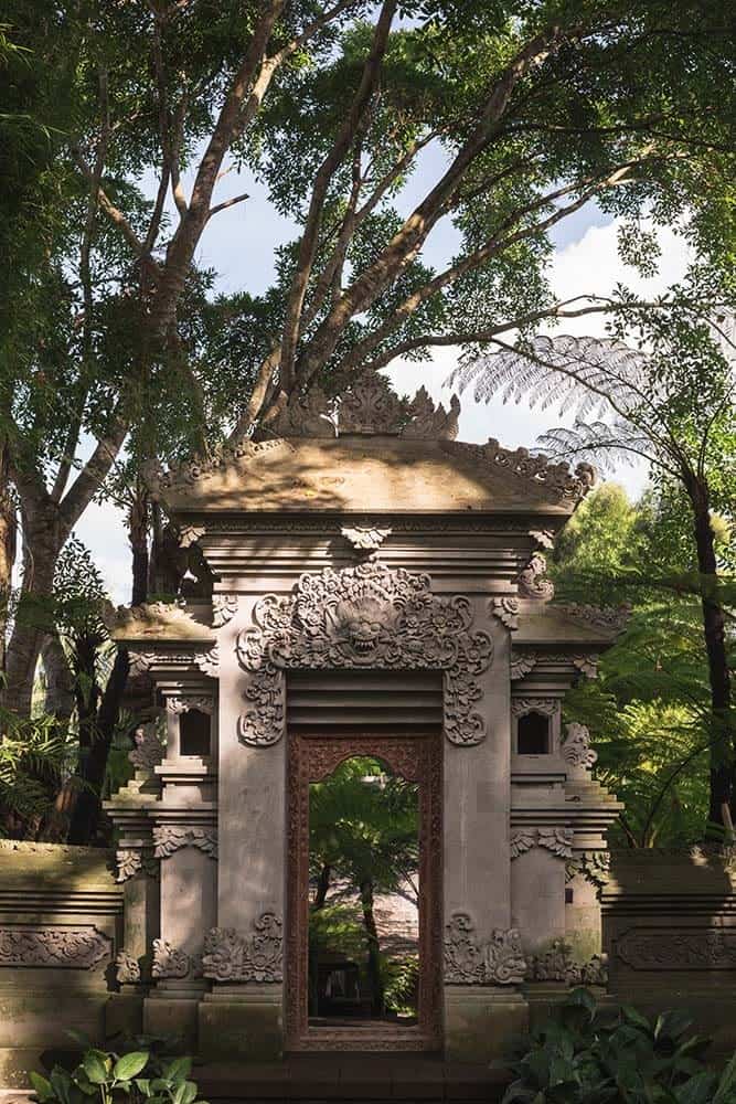 这是房子的大混凝土拱门和外门的全景，上面有复杂的雕刻和图像，使它具有独特的亚洲风格，周围是高大的树木和灌木。