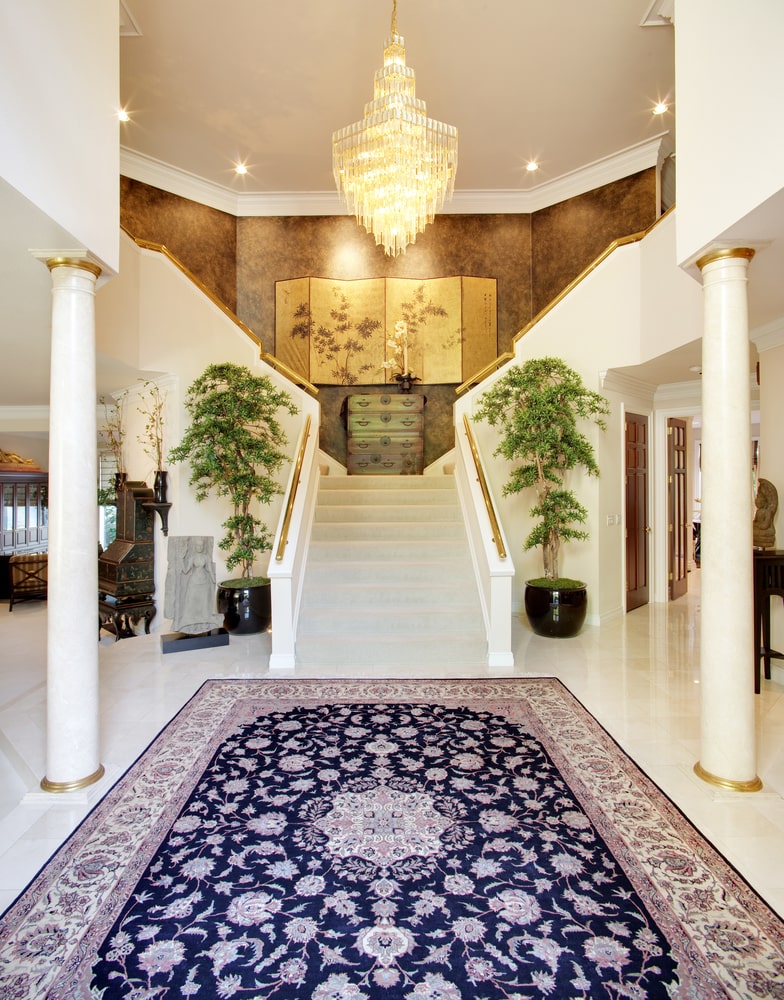 白色楼梯两侧种植着高大的盆栽，楼梯上有金色扶手。这个门厅以大理石柱子和一个大的花地毯为特色，被层叠的水晶吊灯照亮。