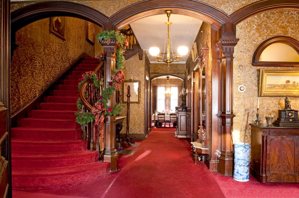 优雅的门厅覆盖着金色图案的壁纸和经典的红地毯，贯穿整个房间。它由一个黄铜吊灯悬挂在木制护墙板包裹的开放式拱门上照亮。