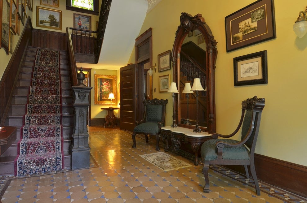 维多利亚风格的门厅与图案瓷砖地板和黄色墙壁充满画廊框架。它拥有经典的楼梯和拱形镜子下的木制控制台桌子，两侧是绿色软垫椅子。