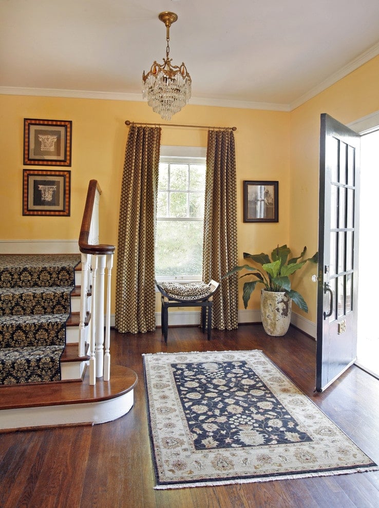 一扇黑色的前门通往门厅，门厅的墙壁是黄色的，窗户上装饰着点状的窗帘。它的特色是一个弯曲的凳子和一个大的花地毯，由层叠的水晶吊灯照亮。