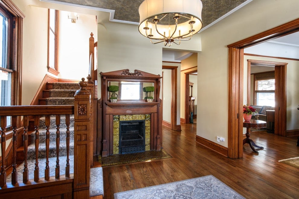 米色的门厅里有一座壁炉和一座木制楼梯，楼梯上装饰着有芙蓉图案的地毯。一盏悬挂在天然硬木地板上的灰色地毯上的鼓形吊灯照亮了房间。
