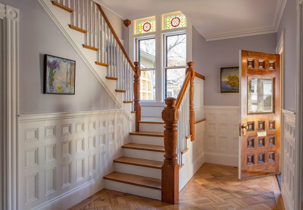 这个门厅展示了一个木制的前门和华丽的艺术品，安装在白色护壁板覆盖的柔软的蓝色墙壁上。它有玻璃窗，浅色硬木地板呈菱形排列。