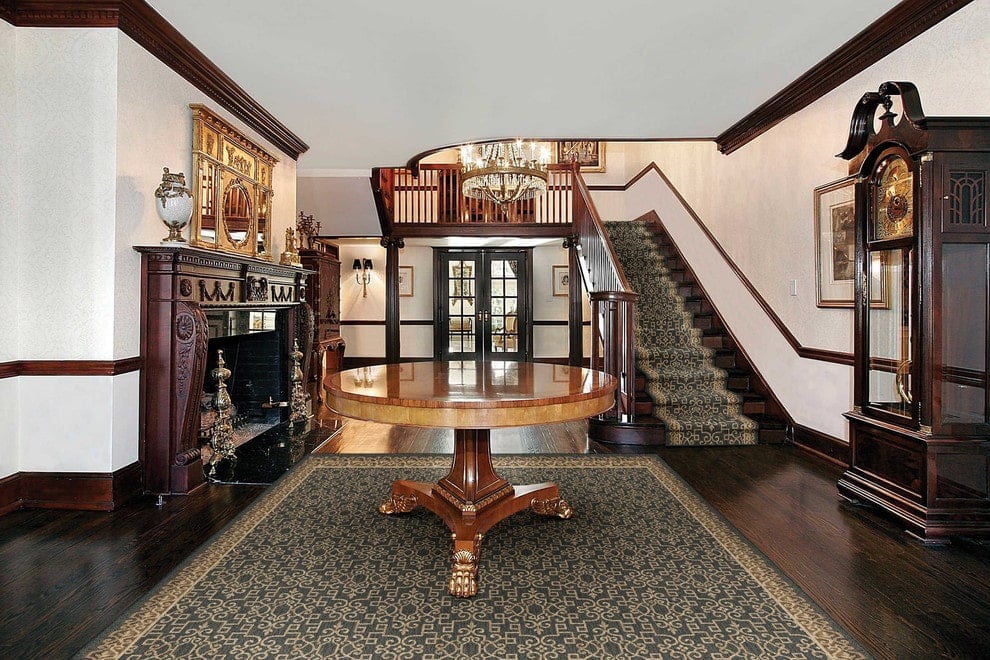 一个有图案的楼梯梯与上面的圆形中心桌子的镶边地毯相匹配。这间门厅拥有一座落地式大座钟和一座带有金色镜子的深色木质壁炉。