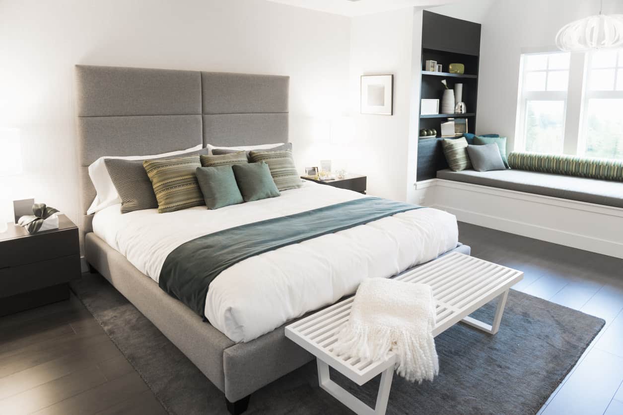 深色宽木板地板与主卧室的原始白色墙壁形成鲜明对比，卧室内有一张灰色的软垫床和一个由独特的枝形吊灯照亮的窗座角落。