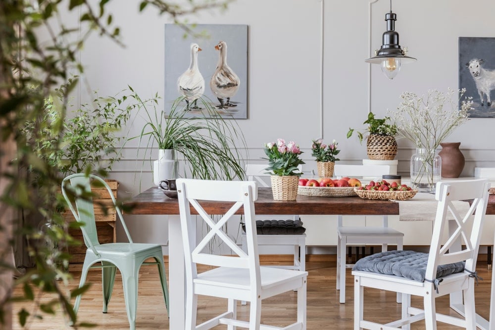 这间舒适的农舍风格的餐厅有漂亮的浅色硬木地板，与白色木腿的木制餐桌很搭配。与此相匹配的是带有绑带靠垫的木制交叉背餐椅。