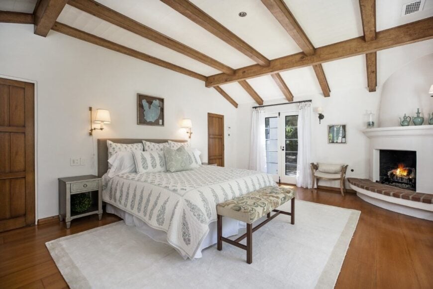 主卧室拥有一张舒适的大床，角落里有一个壁炉。房间以硬木地板和木制横梁的高高的天花板为特色。图片来自Toptenrealestatedeals.com。