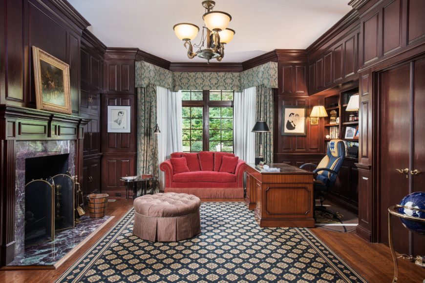 家庭办公室拥有优雅的墙壁和一个优雅的大壁炉。它有一个华丽的办公桌和椅子，还有一个靠近窗户的红色沙发。图片来自Toptenrealestatedeals.com。