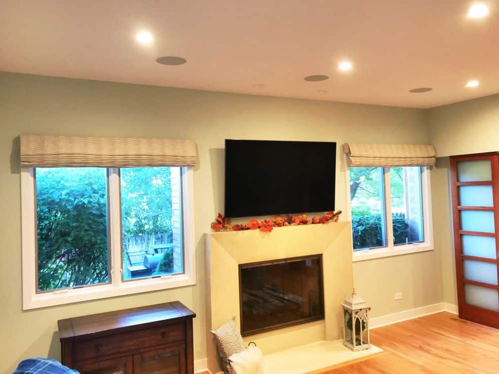 这个起居空间的特色是米黄色的墙壁和硬木地板，以及嵌入式天花板照明。屋内有一座壁炉，墙上有一台平板电视，还有几扇带有罗马风格窗帘的窗户。