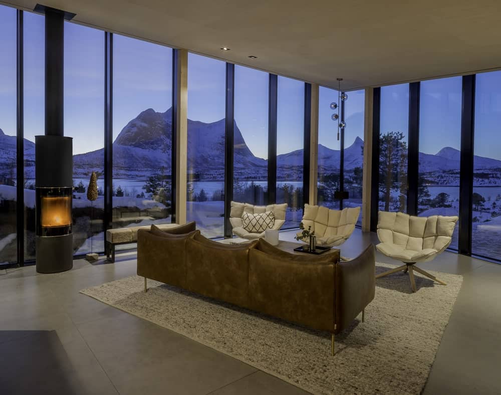 宽敞的斯堪的纳维亚风格的客厅，在挪威北部的小木屋，特色是一个沙发和三把椅子，上面的地毯覆盖着花岗岩瓷砖地板。这个空间被落地玻璃窗包围着。圆柱形的燃木炉就像比利时Stûv公司建造的模型，具有高效的大气温暖设计和360°旋转的玻璃面。一些型号还包括烧烤用的烤架。