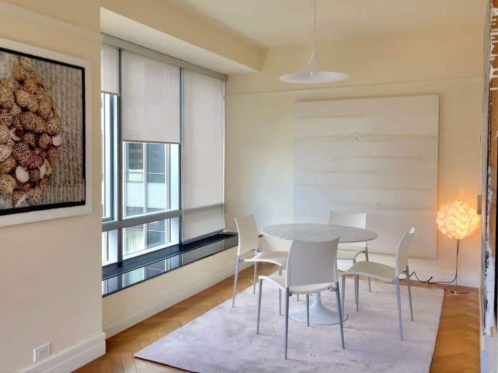 一个漂亮的餐厅，白色的餐桌和椅子设置在覆盖人字形硬木地板的区域地毯上。房间四周是白墙和带遮阳板的玻璃窗。