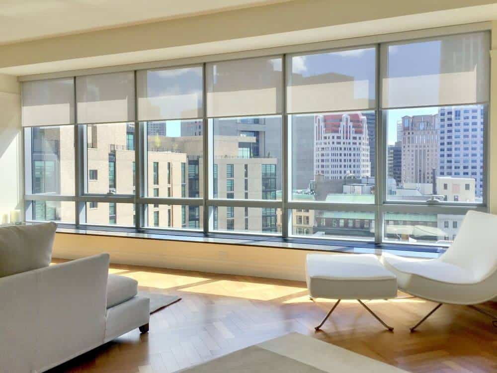 宽敞的客厅拥有人字风格的硬木地板，米黄色的墙壁和时尚的沙发套，以及带有太阳能窗帘的玻璃窗。