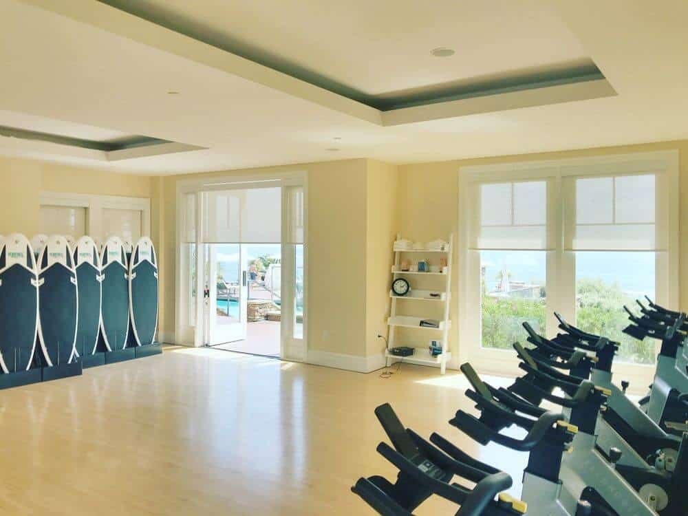 宽敞的家庭健身房，配有抛光的地板和定制的托盘天花板，以及米黄色的墙壁和带太阳能窗帘的窗户。